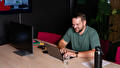 Een man in een groen shirt typt op een laptop aan een houten tafel. Naast hem staan een monitor, een glas water en een zwarte fles.