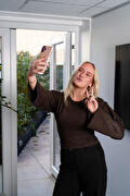 Een vrouw met blond haar houdt haar telefoon omhoog voor een selfie en maakt met haar andere hand een V-teken. Ze staat binnen bij een open glazen deur.