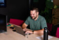 Een man in een groen shirt typt op een laptop aan een houten tafel. Naast hem staan een monitor, een glas water en een zwarte fles.