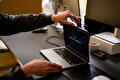 Een persoon wijst naar het scherm van een open laptop met de tekst ‘Google’ op een zoekpagina. Naast de laptop staan een kopje en een telefoon op het bureau.