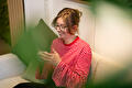 Een glimlachende vrouw met een bril en een rood-wit gestreept shirt zit op een bank en houdt een tablet vast.